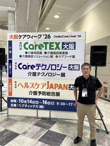 CareTEX大阪’26の会場看板の前に立つ、おむつ宅配便代表の八木大志